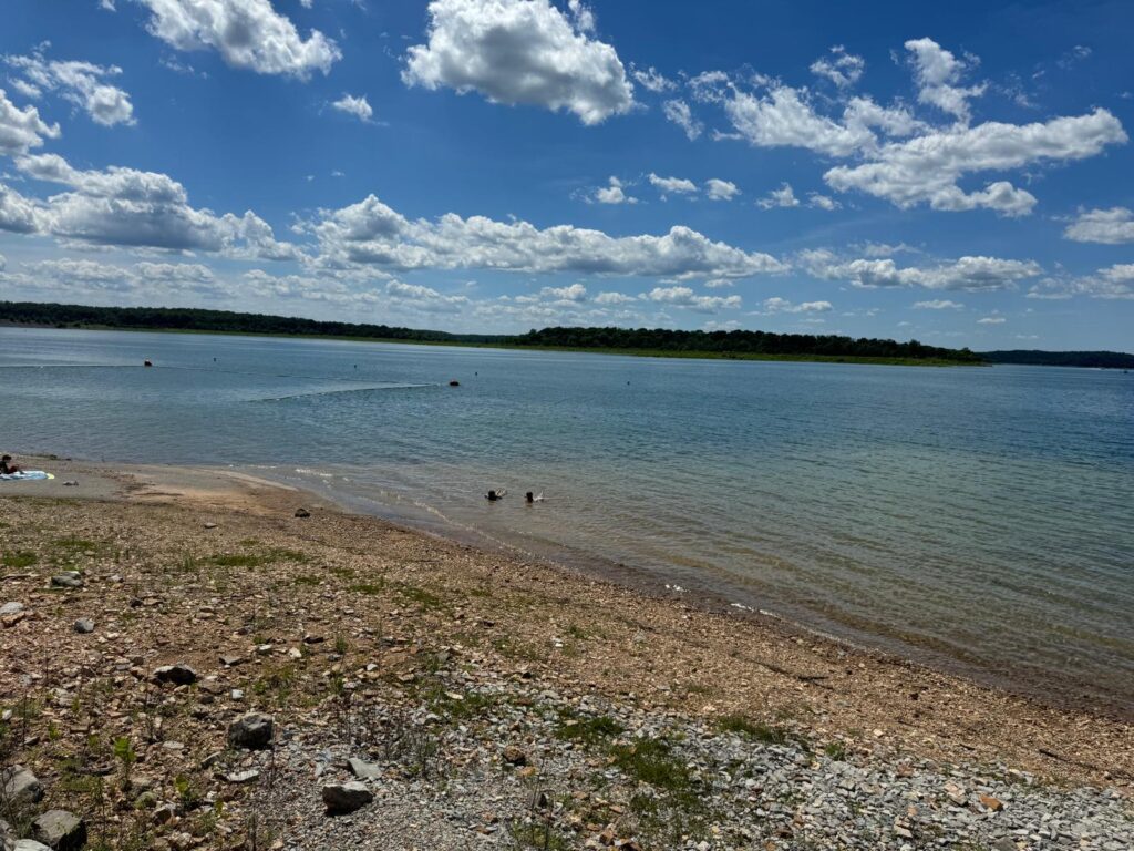 View of a serene lake from a pebble beach under a bright sky with scattered white clouds