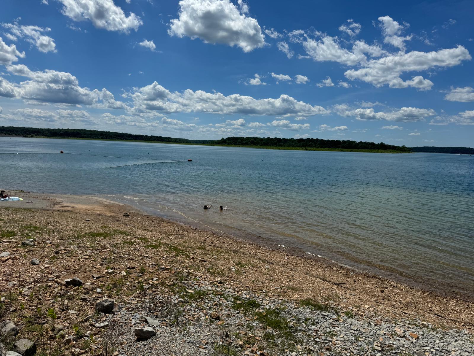 View of a serene lake from a pebble beach under a bright sky with scattered white clouds
